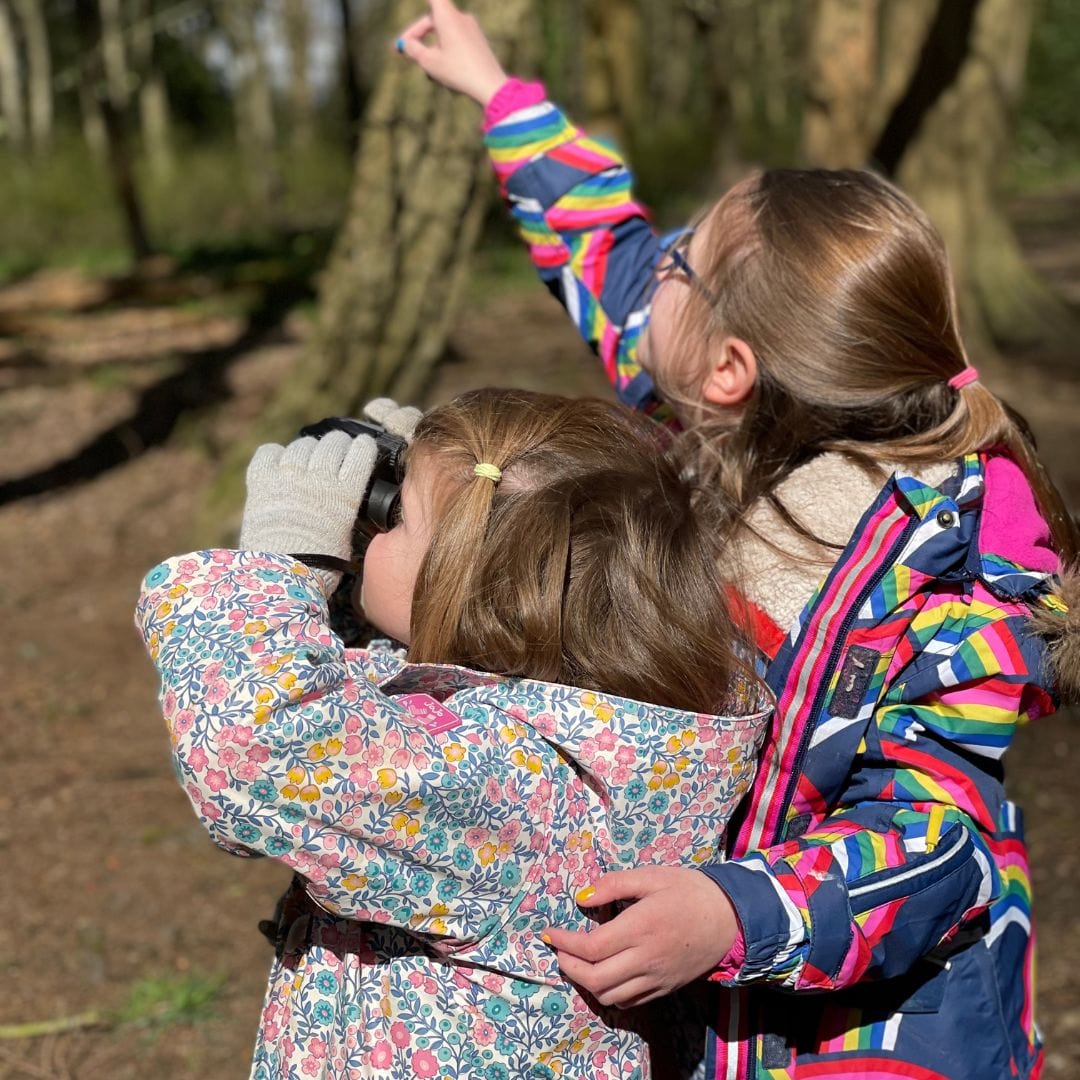 Two young girls looking up into trees through binoculars while birdwatching outdoors, inspired by Little Moments Outside nature spotting book for kids