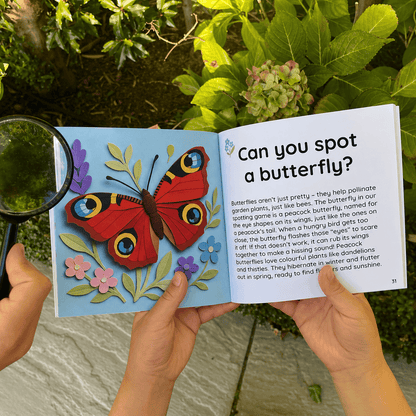 Child's hands holding Little Moments Outside open at the peacock butterfly page in a garden, with a magnifying glass and hydrangea plant in the background